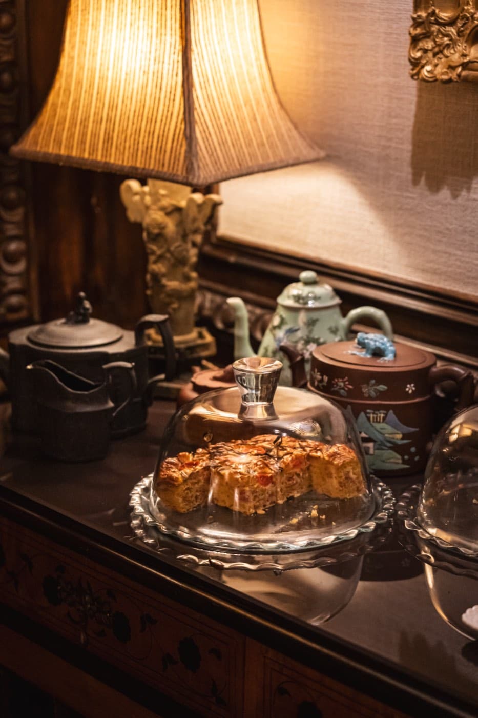 Teapots and a cake under a glass dome on a dark wood sideboard.